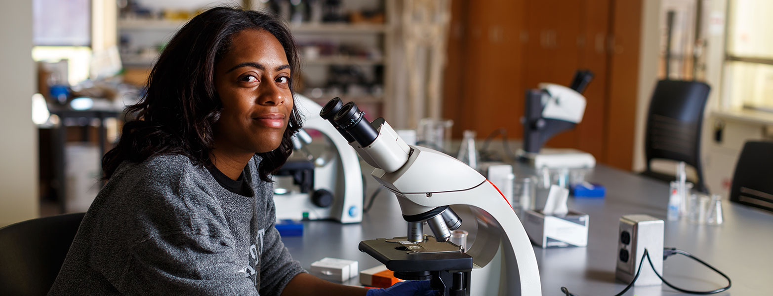 Female student smiles as she looks up from microscope in chemistry lab