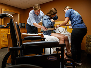 OT professor and student practice assisting a patient moving from a bed to a wheelchair with another student